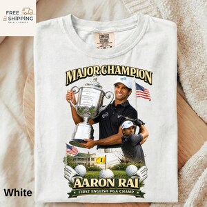 May include: White t-shirt with a golf champion holding a trophy, text reads "MAJOR CHAMPION" and "AARON RAI FIRST ENGLISH PGA CHAMP". The design includes golf balls, a golf course, and the American flag. The shirt is a Comfort Colors brand.