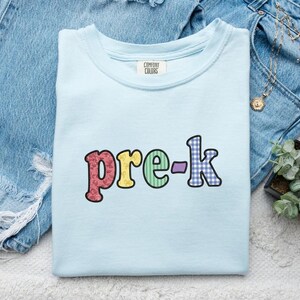 Może przedstawiać: Jasnoniebieska koszulka Comfort Colors z napisem "pre-k" w kolorowych, wzorzystych literach. Litery są obrysowane ciemnobrązowym kolorem. Koszulka jest złożona i umieszczona na niebieskich dżinsach.