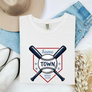 May include: White Comfort Colors t-shirt with a baseball-themed graphic. The design features crossed navy blue baseball bats, a baseball, and the words "home TOWN since 1839" in a red and blue color scheme. The shirt is folded.