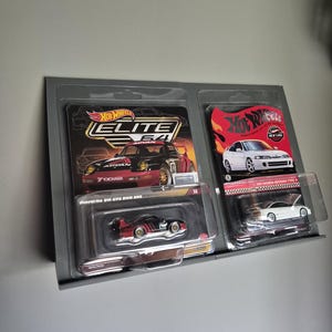 Könnte beinhalten: Zwei Hot Wheels Modellautos in Originalverpackung. Eines ist ein Porsche 911 GT2 EVO 985, das andere ein Honda Integra Type R. Die Autos werden auf einem grauen Regal ausgestellt.