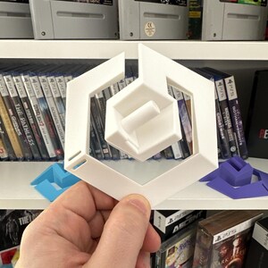 Könnte beinhalten: Ein weißes, geometrisches 3D-gedrucktes Objekt wird vor einem Regal mit Videospielen gehalten. Das Objekt hat einen sechseckigen Außenrahmen mit einer Innenstruktur. Weitere 3D-gedruckte Objekte in Blau und Lila sind ebenfalls sichtbar.
