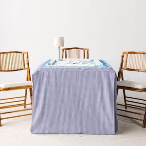 May include: A bamboo game table with a blue and white striped skirt, a light blue lamp, and three bamboo chairs with white cushions. The table has a blue surface with a game board and game pieces.