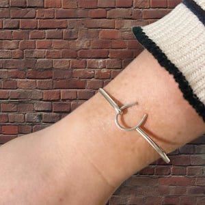 May include: A silver-colored cuff bracelet with a unique hook-and-eye closure. The bracelet is a simple, minimalist design, with a curved hook that fastens to the opposite side. The bracelet is worn on a wrist, against a brick background.