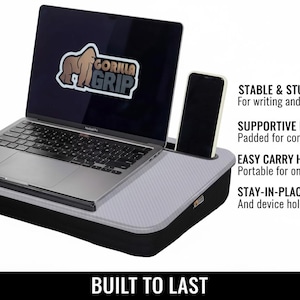Op de afbeelding: Een zwart en grijs laptopbureau met een laptop en een smartphone. De laptop heeft een "Gorilla Grip" logo. Tekst aan de zijkant luidt "Stable & Sturdy", "Supportive Foam", "Easy Carry Handle" en "Stay-in-Place Ledge". De woorden "Built to Last" staan onderaan.