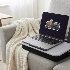 Op de afbeelding: Een laptop met het "GORILLA GRIP" logo op het scherm staat op een zwart en grijs laptopbureau met een telefoonhouder. Het bureau staat op een grijze bank met een cr&egrave;mekleurige deken.