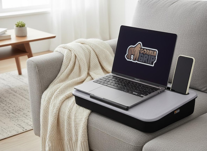 Op de afbeelding: Een laptop met het "GORILLA GRIP" logo op het scherm staat op een zwart en grijs laptopbureau met een telefoonhouder. Het bureau staat op een grijze bank met een cr&egrave;mekleurige deken.