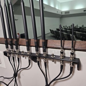 Pu&ograve; includere: Un supporto in metallo nero montato a parete che sostiene diverse antenne wireless nere con connettori BNC argentati. Le antenne sono disposte in fila, con cavi neri che si estendono verso il basso da ciascun connettore verso un punto centrale.
