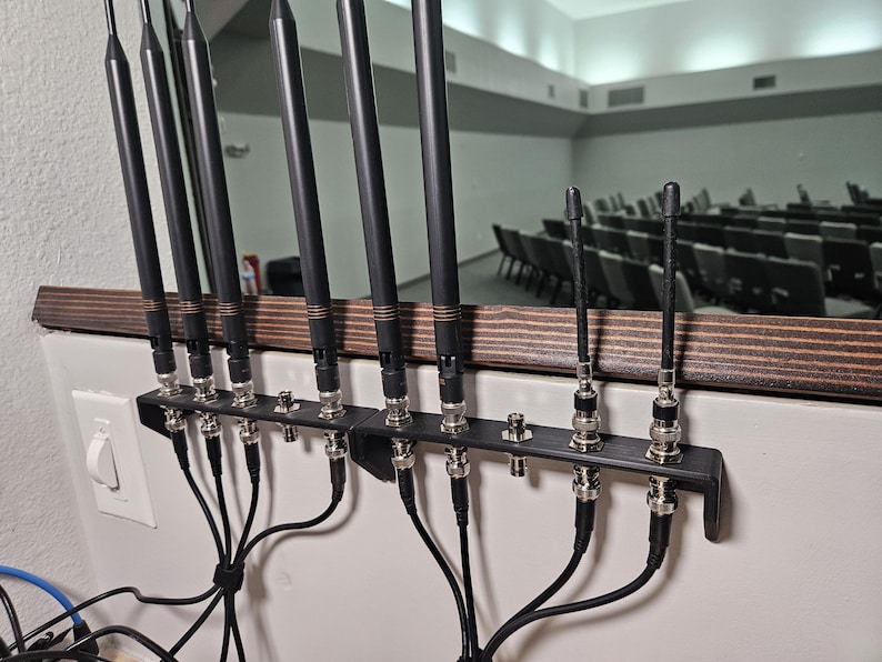 Pu&ograve; includere: Un supporto in metallo nero montato a parete che sostiene diverse antenne wireless nere con connettori BNC argentati. Le antenne sono disposte in fila, con cavi neri che si estendono verso il basso da ciascun connettore verso un punto centrale.