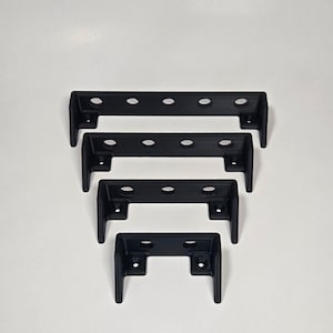 Pu&ograve; includere: Quattro staffe metalliche a forma di U di colore nero opaco disposte in ordine decrescente di dimensioni. Ogni staffa presenta una barra orizzontale con fori circolari e due gambe rivolte verso il basso, ciascuna con un foro per vite.