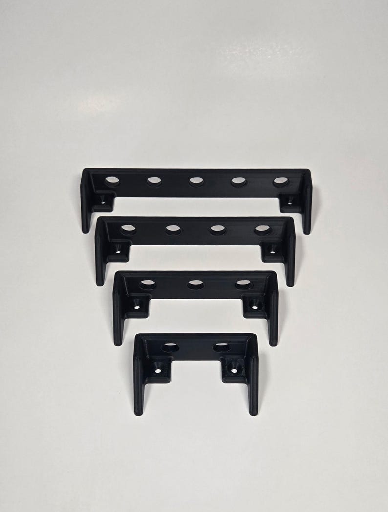 Pu&ograve; includere: Quattro staffe metalliche a forma di U di colore nero opaco disposte in ordine decrescente di dimensioni. Ogni staffa presenta una barra orizzontale con fori circolari e due gambe rivolte verso il basso, ciascuna con un foro per vite.