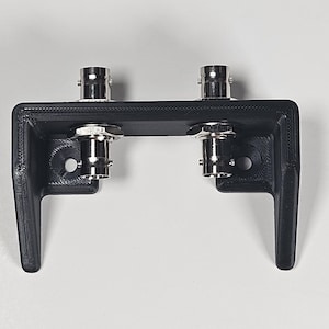 Pu&ograve; includere: Una staffa di montaggio nera stampata in 3D con due connettori BNC metallici installati sulla superficie orizzontale superiore. La staffa ha un design a forma di U con due gambe rivolte verso il basso e due fori di montaggio circolari.