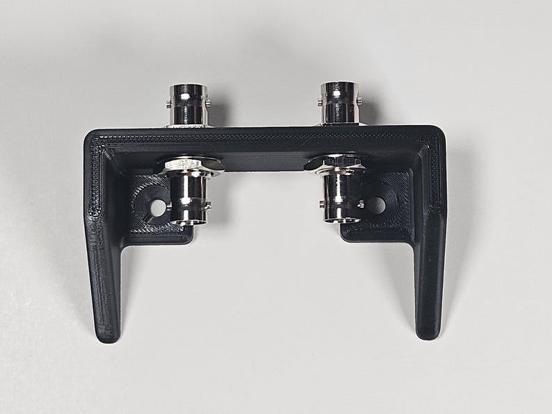 Pu&ograve; includere: Una staffa di montaggio nera stampata in 3D con due connettori BNC metallici installati sulla superficie orizzontale superiore. La staffa ha un design a forma di U con due gambe rivolte verso il basso e due fori di montaggio circolari.