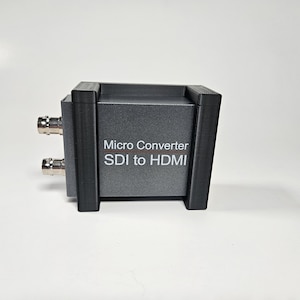 May include: A gray Micro Converter SDI to HDMI device held within a black 3D-printed mounting bracket. The unit features two silver BNC connectors on the side and white text printed on the front face identifying the converter model.