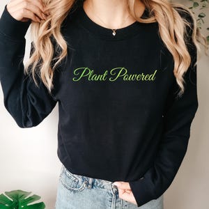 Könnte beinhalten: Schwarzes Langarm-Sweatshirt mit dem grünen Schriftzug "Plant Powered". Das Sweatshirt wird mit Jeansshorts kombiniert. Das Bild vermittelt einen entspannten, lässigen Stil.