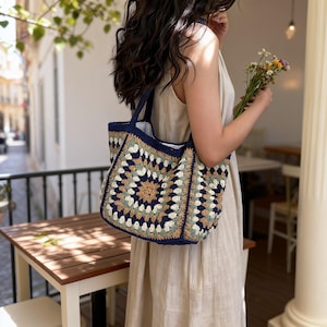 May include: A handmade crochet tote bag with a granny square design. The bag features a navy blue border and handles, with a central pattern in shades of beige, green, and cream. The bag is held by a person wearing a beige dress.