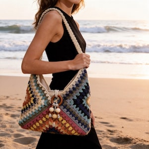 May include: A colorful, crocheted tote bag with a geometric pattern. The bag features a cream-colored braided strap and a wooden ring with decorative beads. The bag's design incorporates various colors, including blue, pink, orange, and cream.