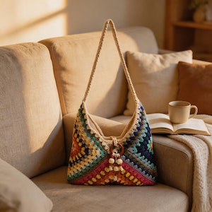 May include: A colorful, handmade crochet bag with a beige strap and wooden ring detail. The bag features a geometric pattern in shades of blue, green, yellow, pink, and orange. The bag is sitting on a beige couch.