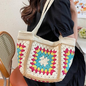 May include: A cream-colored crochet tote bag with a colorful square pattern. The square pattern features a central blue square surrounded by yellow and pink squares. The bag has cream-colored straps and a brown border.