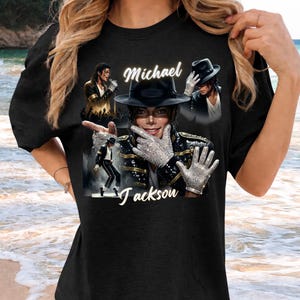 May include: Black t-shirt featuring multiple images of Michael Jackson. The design includes the name "Michael Jackson" in white script, along with images of the singer in various poses, wearing a hat and gloves.