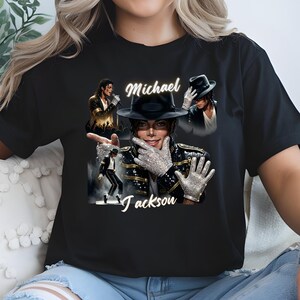 May include: Black t-shirt featuring multiple images of Michael Jackson, including a portrait with a black hat and sparkling gloves. The name "Michael Jackson" is written in a stylized font across the shirt. The shirt is a classic crew neck.