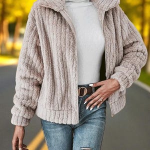 May include: A beige, textured zip-up jacket with a wide collar. The jacket has long sleeves and is paired with a white turtleneck and blue jeans. A brown belt with a gold buckle is visible.