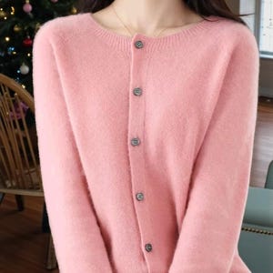 Puede incluir: Un cárdigan rosa claro con cuello redondo y mangas largas. Tiene una botonadura con cuatro botones pequeños y redondos. El material parece suave y el estilo general es informal.