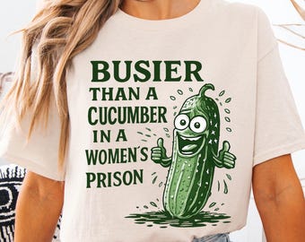 May include: Cream-colored t-shirt with the text "BUSIER THAN A CUCUMBER IN A WOMEN'S PRISON" and a cartoon cucumber giving a thumbs-up. The graphic is in green and black.