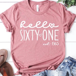 May include: A dusty rose t-shirt with the words "hello SIXTY ONE est. 1965" in white script. The shirt is paired with blue jeans and brown sandals. A small potted plant is also in the image.