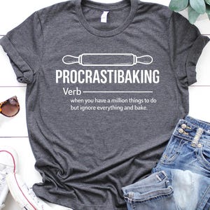 May include: Dark gray t-shirt with the text "PROCRASTIBAKING" and a rolling pin graphic. The definition reads "Verb: when you have a million things to do but ignore everything and bake." The shirt is paired with blue jeans and white sneakers.