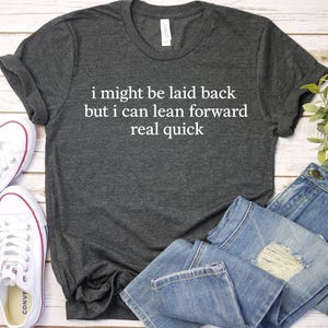 May include: Dark gray t-shirt with the text "i might be laid back but i can lean forward real quick." The image also shows white sneakers, and blue jeans.