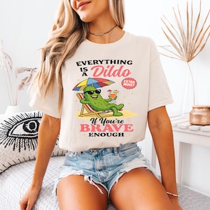 May include: Cream-colored t-shirt with the text "Everything is a Dildo If You're Brave Enough" and a cartoon pickle character relaxing on a beach chair. The pickle wears sunglasses and holds a cocktail under a rainbow umbrella.
