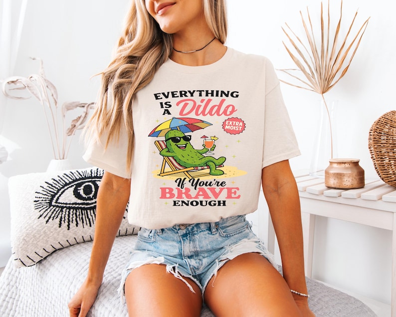 May include: Cream-colored t-shirt with the text "Everything is a Dildo If You're Brave Enough" and a cartoon pickle character relaxing on a beach chair. The pickle wears sunglasses and holds a cocktail under a rainbow umbrella.