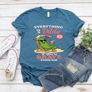 May include: Teal t-shirt with the text "Everything is a Dildo" and a cartoon pickle wearing sunglasses, relaxing on a beach chair with a cocktail. The shirt also features the text "If You're Brave Enough". White sneakers and blue jeans are also in the image.