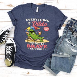 May include: Navy blue t-shirt with the text "Everything is a Dildo" and a cartoon pickle character relaxing on a beach chair. The shirt is paired with blue jeans and black sneakers.