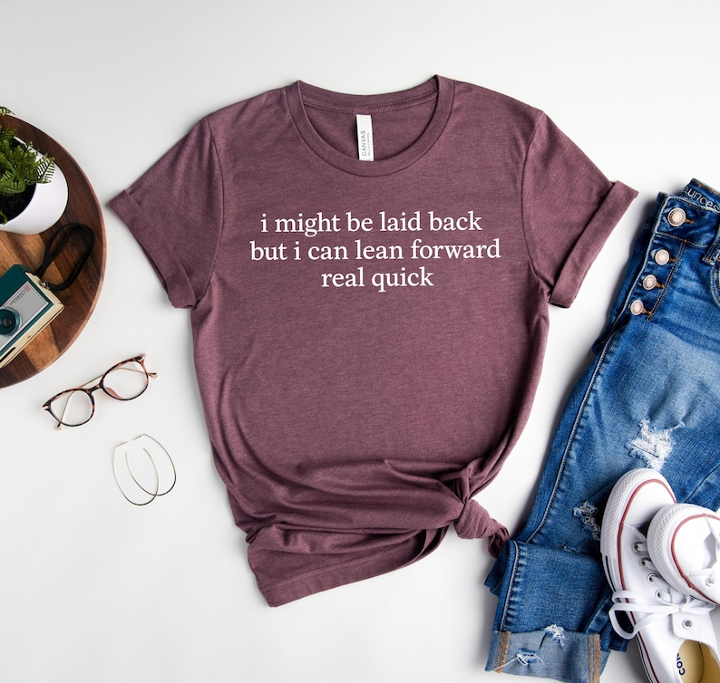 May include: A maroon t-shirt with the text "i might be laid back but i can lean forward real quick" in white. The shirt is paired with blue jeans and white sneakers. Other accessories include glasses and earrings.