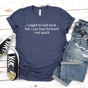 May include: A navy blue t-shirt with the text "i might be laid back but i can lean forward real quick" in white. The shirt is paired with blue jeans, a watch, and black and white sneakers.