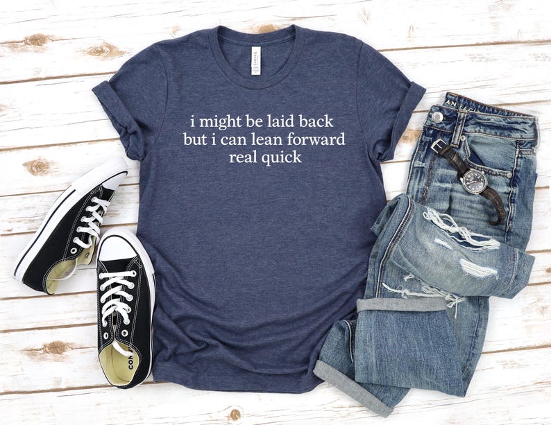 May include: A navy blue t-shirt with the text "i might be laid back but i can lean forward real quick" in white. The shirt is paired with blue jeans, a watch, and black and white sneakers.