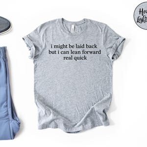 May include: Heather light grey t-shirt with the text "i might be laid back but i can lean forward real quick". The shirt is paired with blue athletic pants and a gray sneaker.