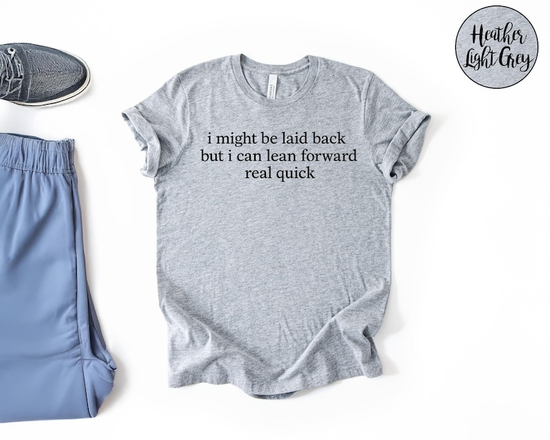 May include: Heather light grey t-shirt with the text "i might be laid back but i can lean forward real quick". The shirt is paired with blue athletic pants and a gray sneaker.