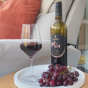 May include: A bottle of Freixenet Mía red wine with a black label, a clear glass filled with red wine, and a cluster of fresh red grapes arranged on a round white tray. The wine bottle features the text Mía, Barcelona, and 2023.