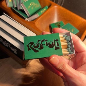 Puede incluir: Una caja de cerillas rectangular con una cubierta verde que presenta el texto negro Roscioli. La caja está parcialmente abierta, revelando una fila de cerillas de madera con puntas de color verde oscuro. Otras cajas idénticas son visibles.