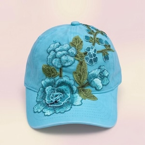 Puede incluir: Una gorra de béisbol azul claro con un bordado intrincado de grandes flores color verde azulado y hojas verdes que se extienden desde el lado inferior izquierdo hacia la corona. La gorra tiene una visera curva y un botón a juego en la parte superior.