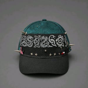 May include: A dark green and black baseball cap featuring a central band with a grey Celtic dragon knotwork pattern. The cap is embellished with silver-toned metal spikes, small star-shaped studs, a miniature red and white pill charm, and a pink tag.