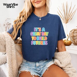May include: Navy blue t-shirt with the text "IT'S A GREAT DAY TO BUILD FUTURES" in colorful letters. The shirt is a casual style, suitable for everyday wear.