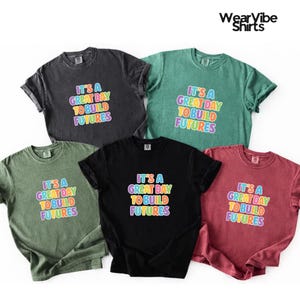 May include: Five short-sleeved t-shirts in various colors: black, dark gray, green, red, and olive green. Each shirt features the text "IT'S A GREAT DAY TO BUILD FUTURES" in colorful, bubble-style letters. The brand name "WearVibe Shirts" is in the top right corner.