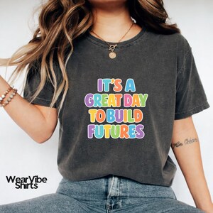 May include: Dark gray t-shirt with the text "IT'S A GREAT DAY TO BUILD FUTURES" in colorful bubble letters. The shirt has a crew neck and short sleeves. The WearVibe Shirts logo is visible in the bottom left corner.