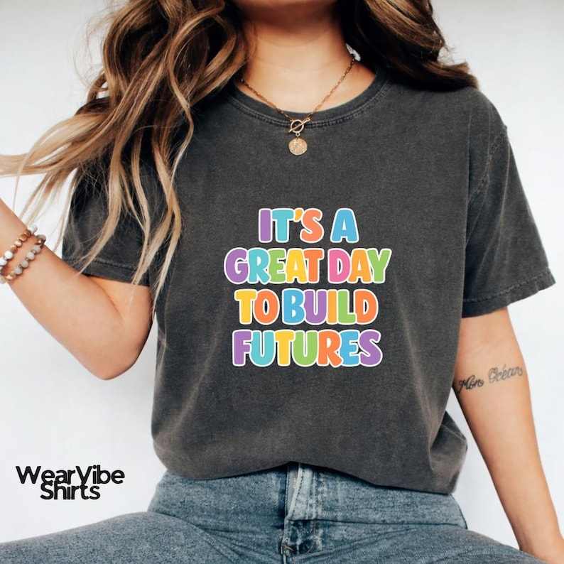 May include: Dark gray t-shirt with the text "IT'S A GREAT DAY TO BUILD FUTURES" in colorful bubble letters. The shirt has a crew neck and short sleeves. The WearVibe Shirts logo is visible in the bottom left corner.