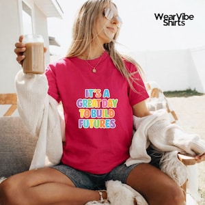 May include: A magenta t-shirt with the text "IT'S A GREAT DAY TO BUILD FUTURES" in rainbow colors. The shirt is being worn by a person holding a glass of iced coffee.