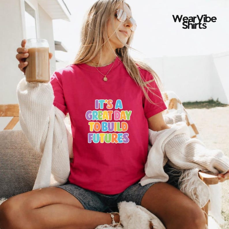 May include: A magenta t-shirt with the text "IT'S A GREAT DAY TO BUILD FUTURES" in rainbow colors. The shirt is being worn by a person holding a glass of iced coffee.