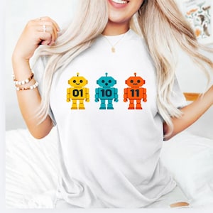 May include: White t-shirt with three robot designs in yellow, teal, and orange. Each robot has a different number: 01, 10, and 11. The robots have a simple, geometric design.